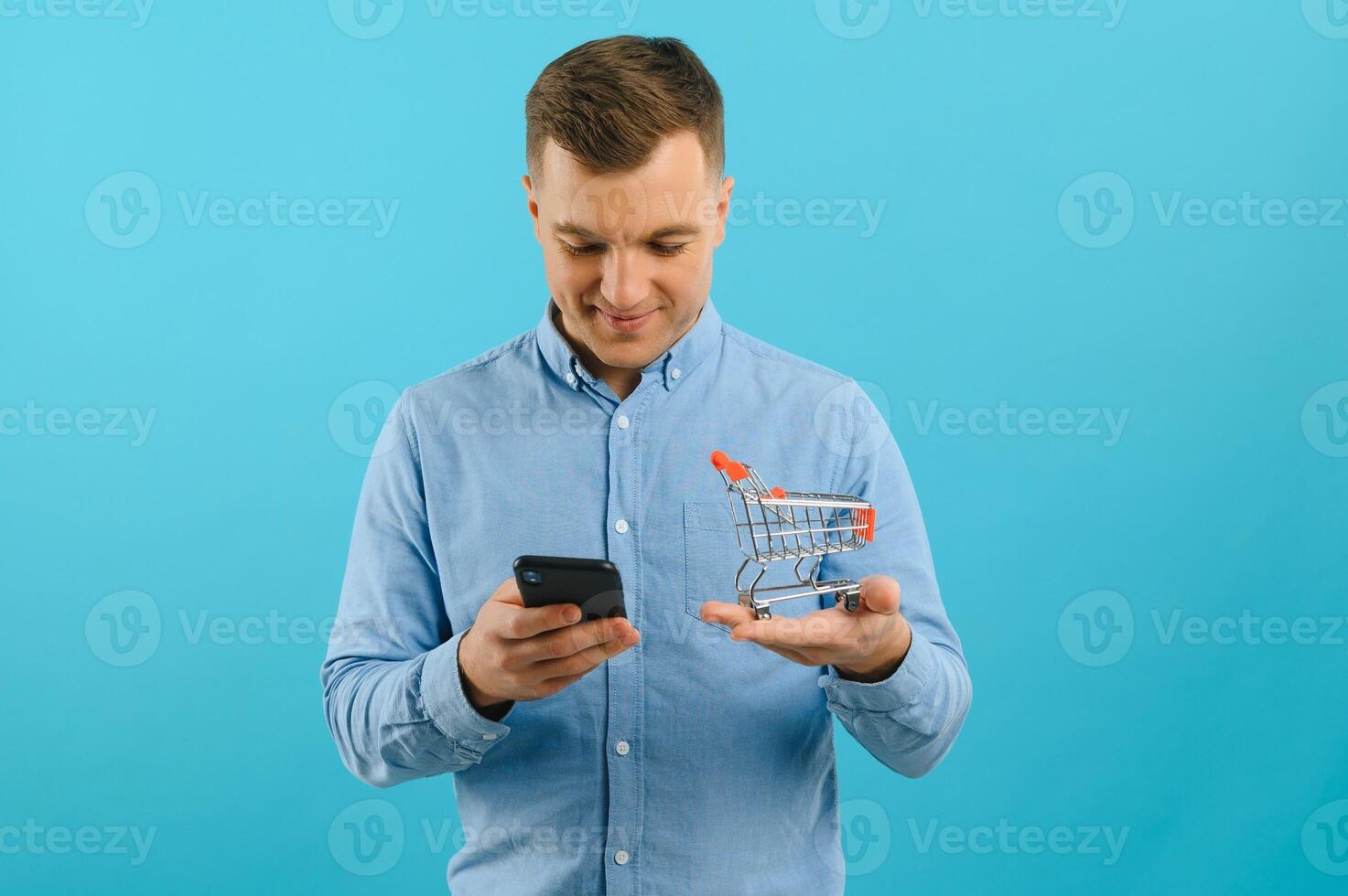 A young man holding a mini shopping cart and standing in front of a blue background in the studio. Sales concept. Place for text. photo