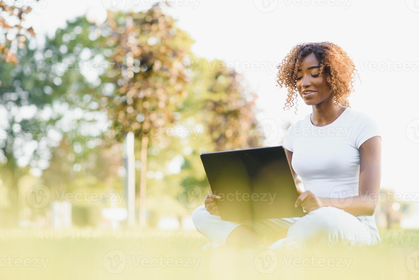 Young attractive dark-skinned college student wearing tank top and jeans sitting on the lawn at campus on sunny day, working on her thesis using laptop computer, looking busy and concentrated photo