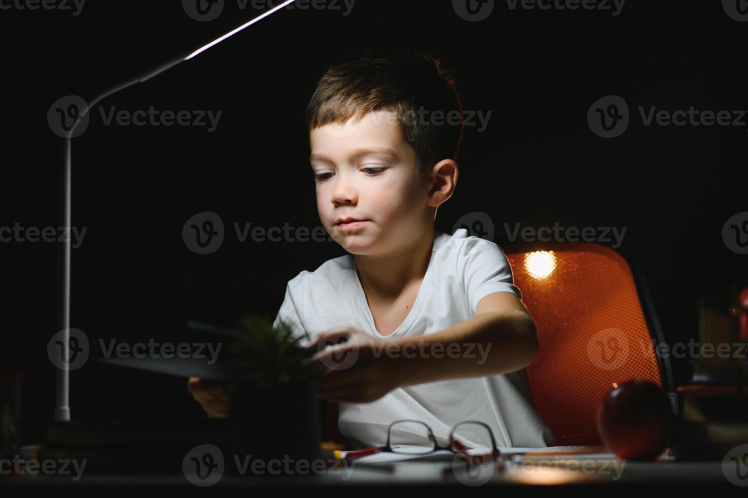 concentrated schoolboy reading book at table with books, plant, lamp, colour pencils, apple, and textbook photo
