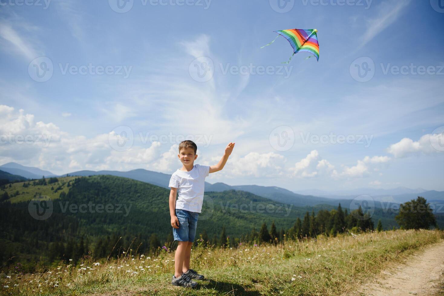 Little school age boy running down the slope with kite in the sun. Sunny summer or spring day at sunset. Active outdoor games and leisure. Mountain landscape with hills and coniferous trees. photo