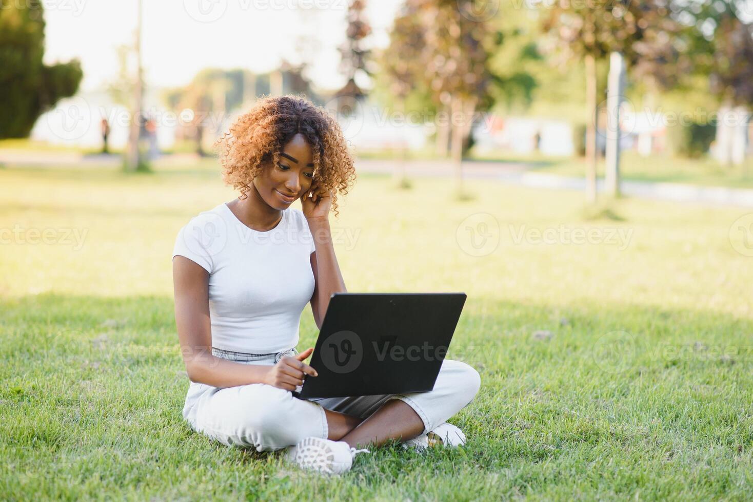 Young attractive dark-skinned college student wearing tank top and jeans sitting on the lawn at campus on sunny day, working on her thesis using laptop computer, looking busy and concentrated photo