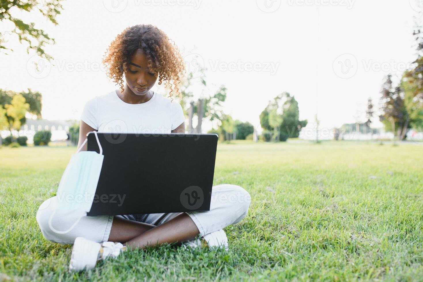Thoughtful cute mixed female international student with curly hair is sitting on fresh grass with modern laptop in public park, leaning on apple tree and wistfully looking aside during her break photo
