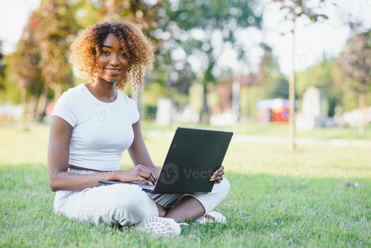 Thoughtful cute mixed female international student with curly hair is sitting on fresh grass with modern laptop in public park, leaning on apple tree and wistfully looking aside during her break photo