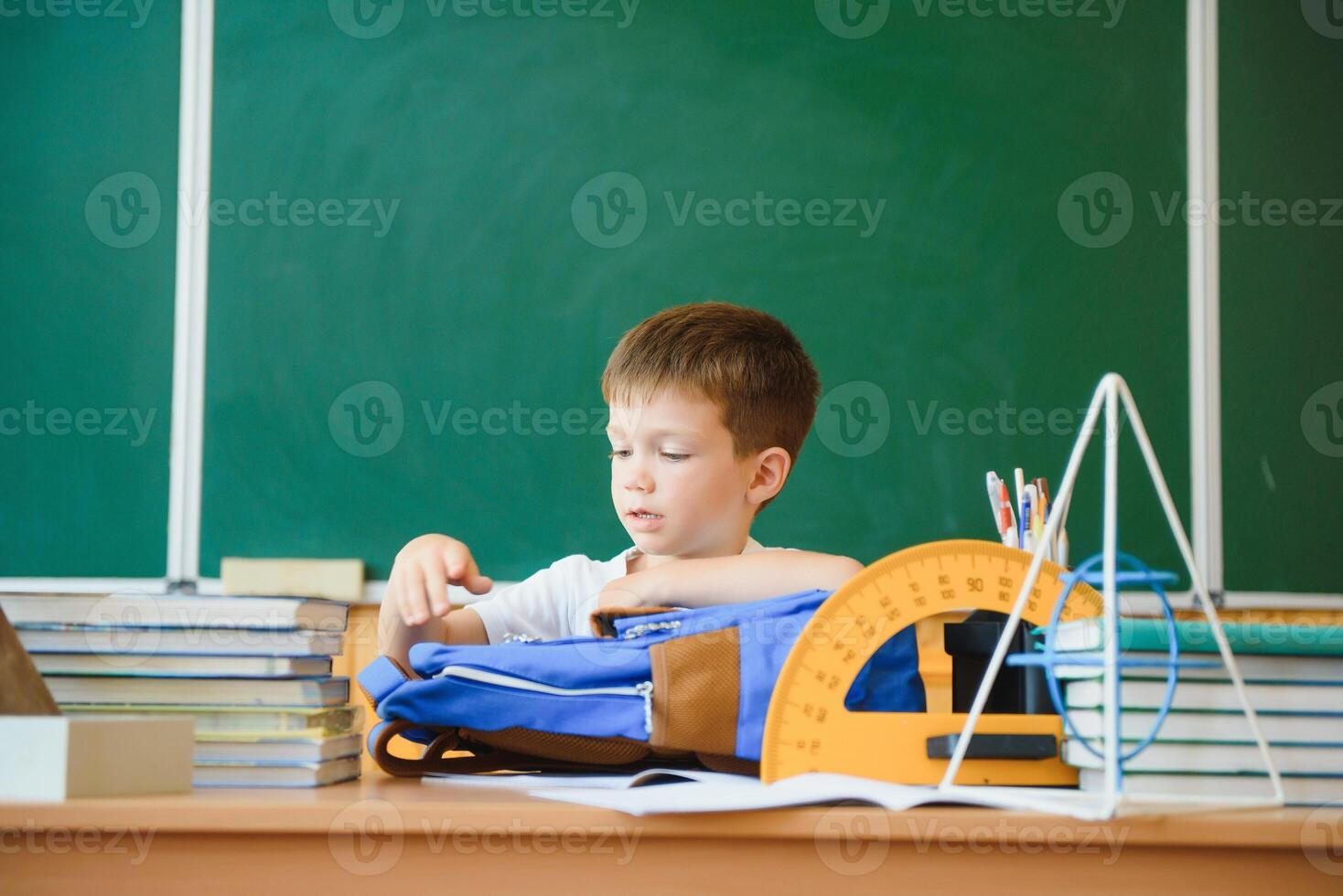 Happy cute clever boy is sitting at a desk. Child is ready to answer with a blackboard on a background. Ready for school. Back to school. books on desk photo