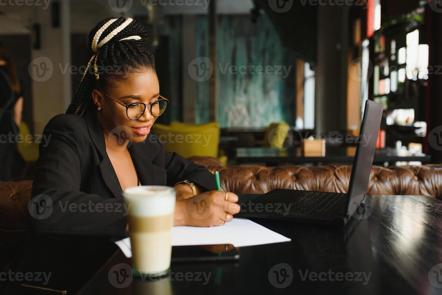 estudiante niña hojeada Internet, utilizando gratis Wifi a cafetería. africano persona de libre dedicación pensando en ideas para su Blog, utilizando ordenador portátil a trabajo colaborativo espacio, descansando mano en de madera mesa, mirando con inspirado sonrisa foto