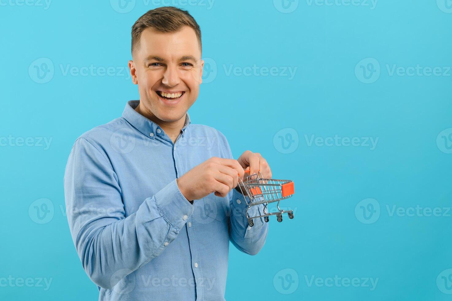 Young smiling man holding a mini shopping cart on a blue background. Sales concept. Place for text. photo