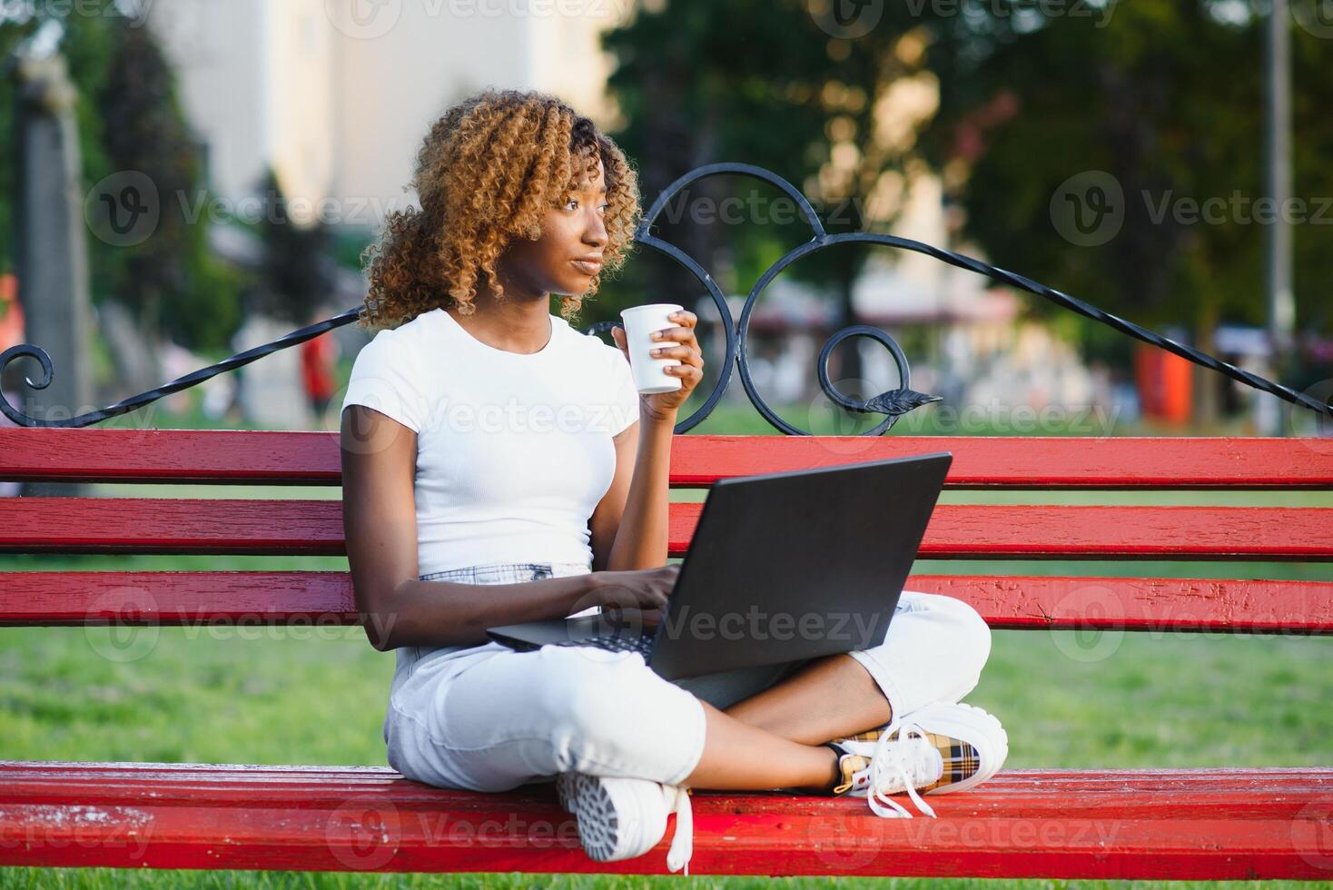 Pretty African American girl sitting on bench and talking on cellphone while working on laptop sitting at table with coffee on street photo