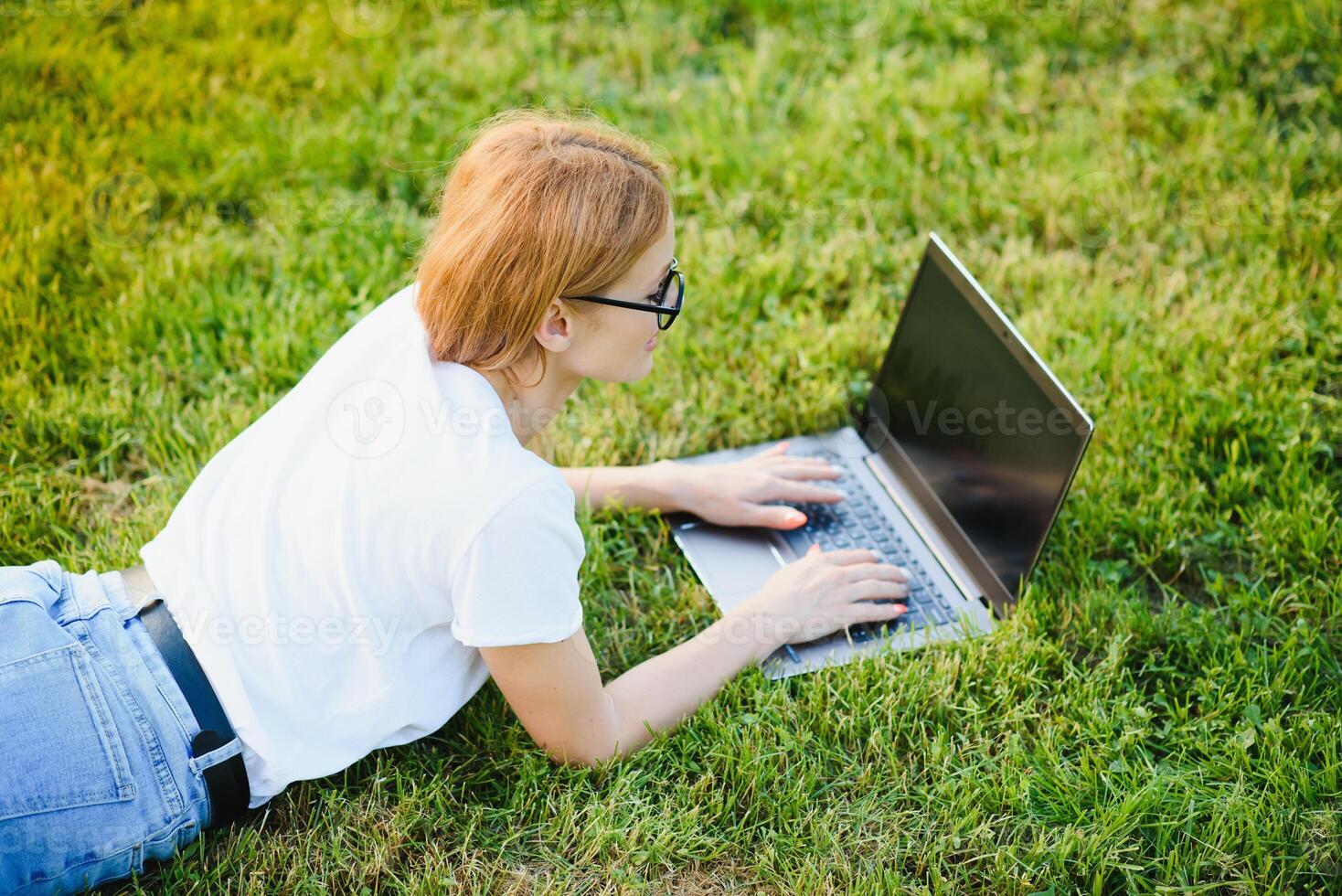 Happy middle aged woman lying on green grass using laptop computer in the park photo