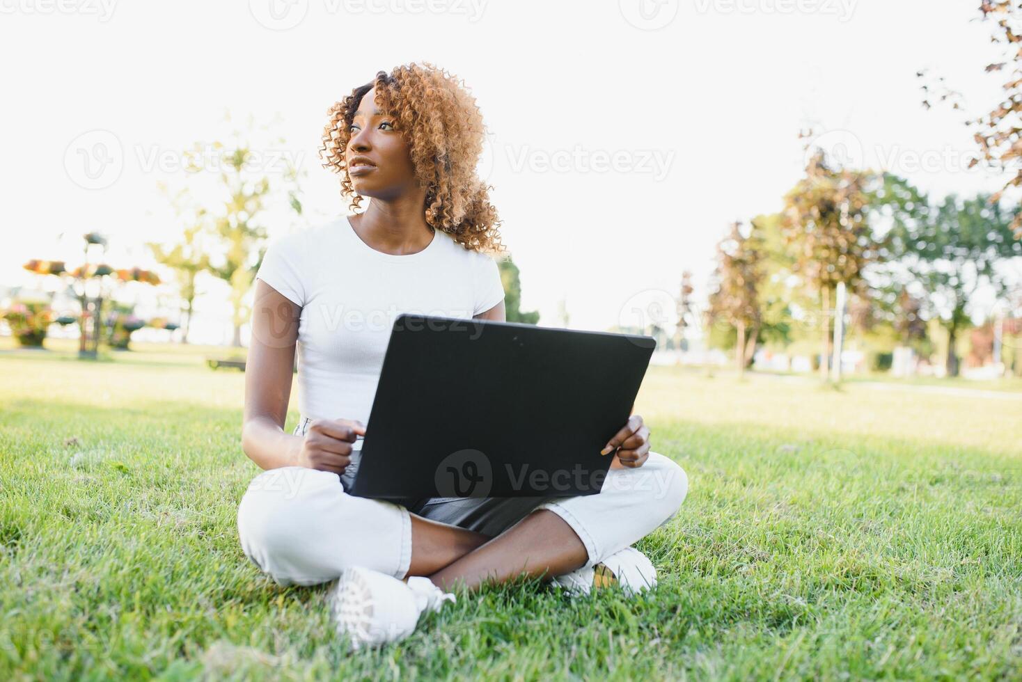 pensativo linda mezclado hembra internacional estudiante con Rizado pelo es sentado en Fresco césped con moderno ordenador portátil en público parque, propensión en manzana árbol y con nostalgia mirando aparte durante su descanso foto