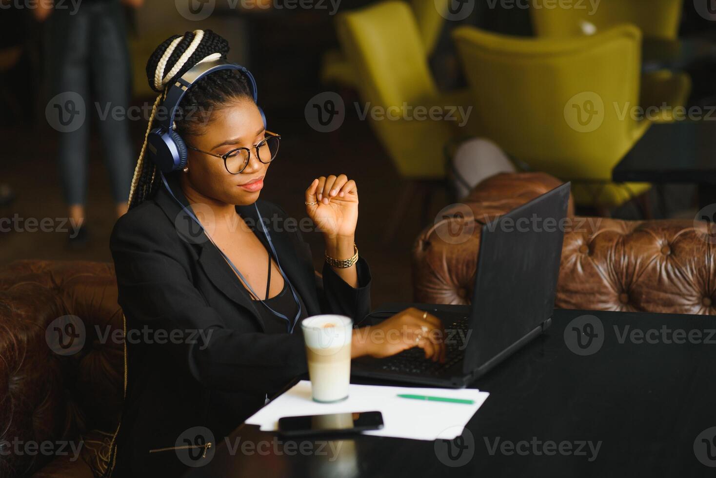 Happy black girl in wireless headphones studying online, using laptop and taking notes, cafe interior, copy space photo