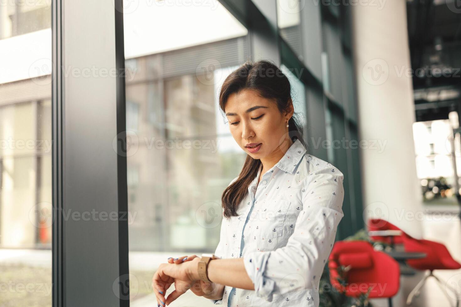 enfocado mujer de negocios mirando en su muñeca reloj mientras en pie cerca oficina ventana foto
