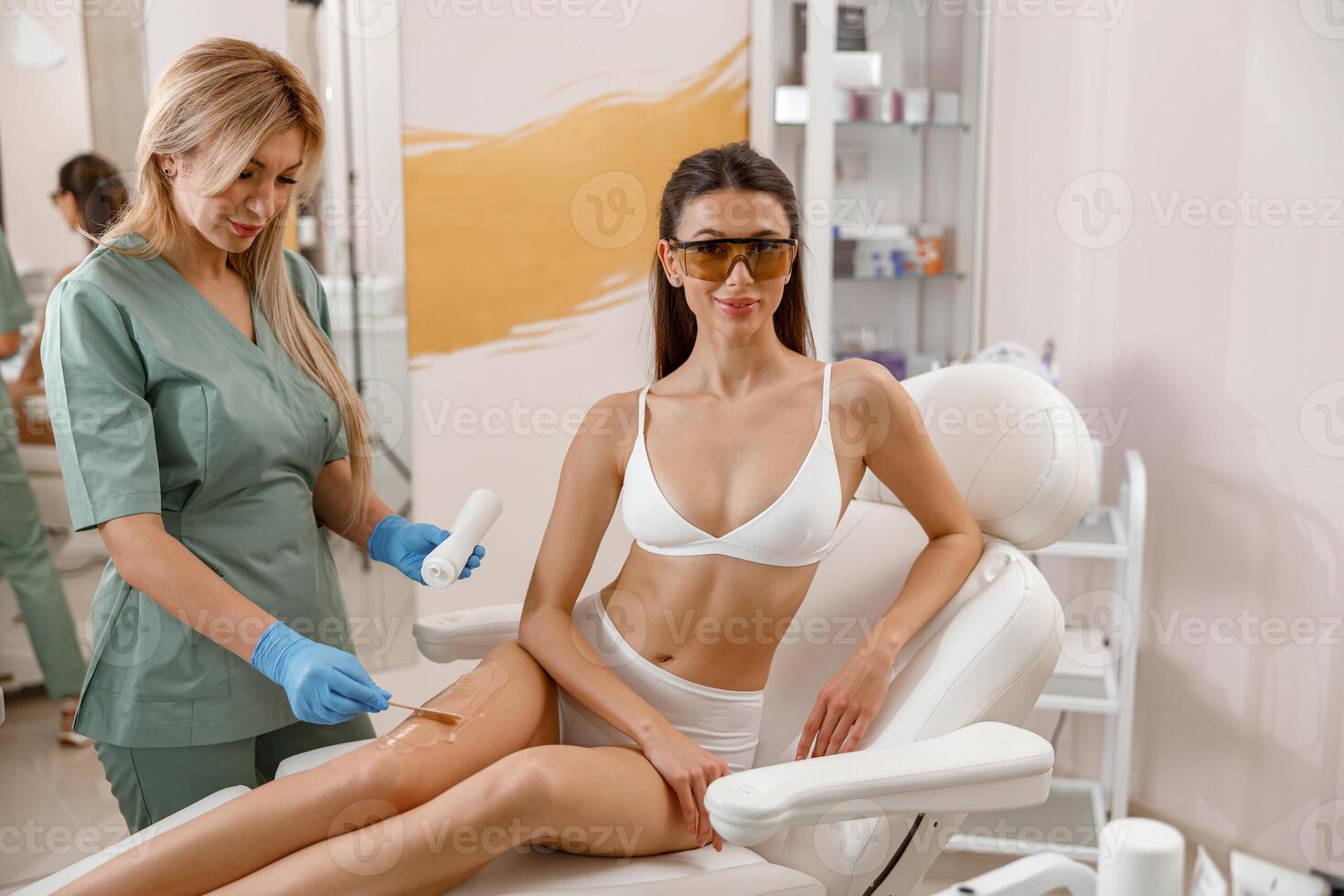 Cosmetologist applying ointment on woman legs before hair removal procedure in beauty salon photo