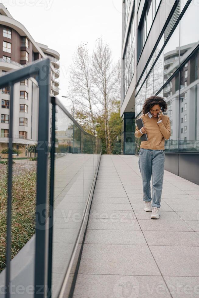 Female freelancer is talking phone while standing with laptop on modern building background photo