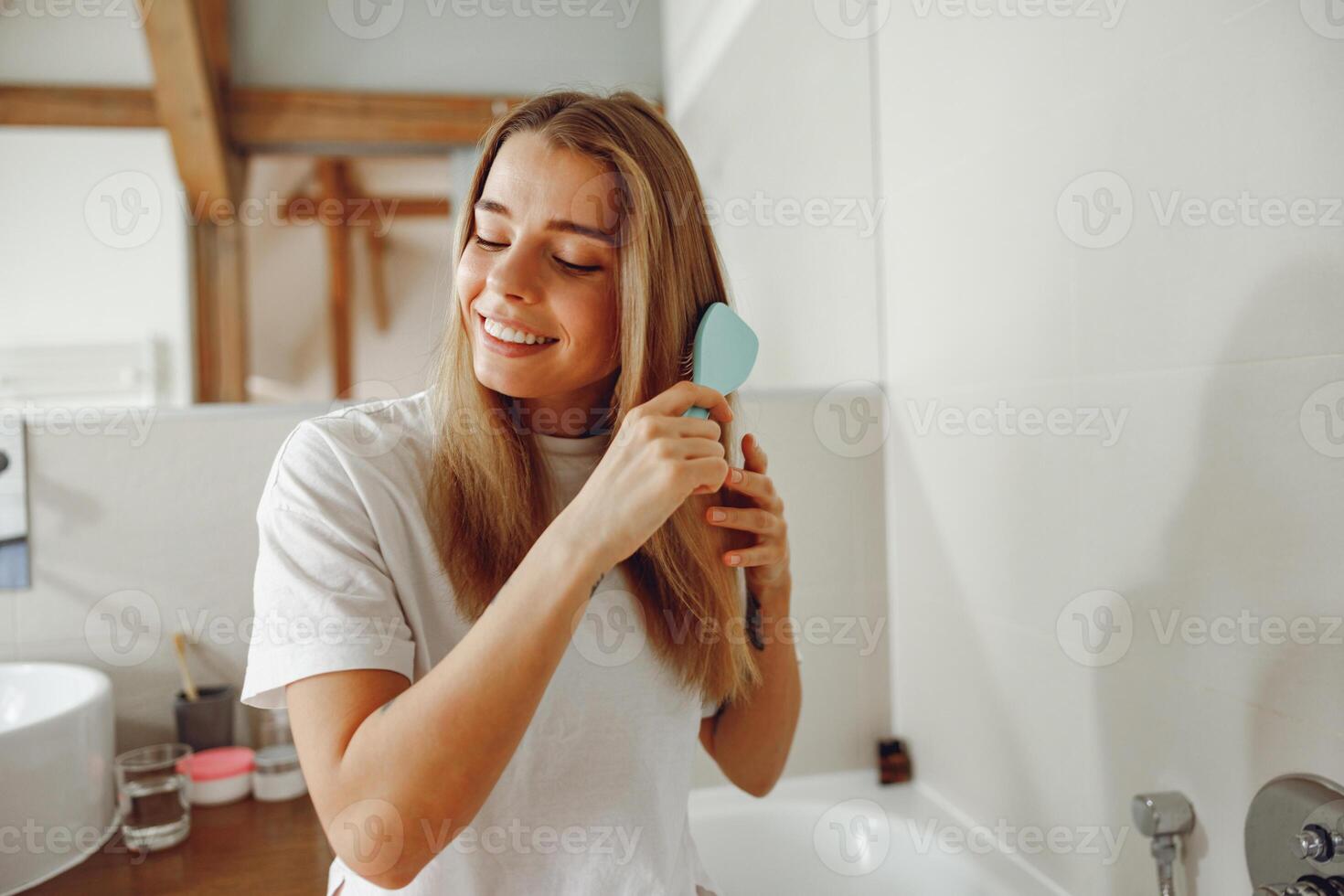 Young woman brushing her hair with comb while standing in bathroom near mirror photo