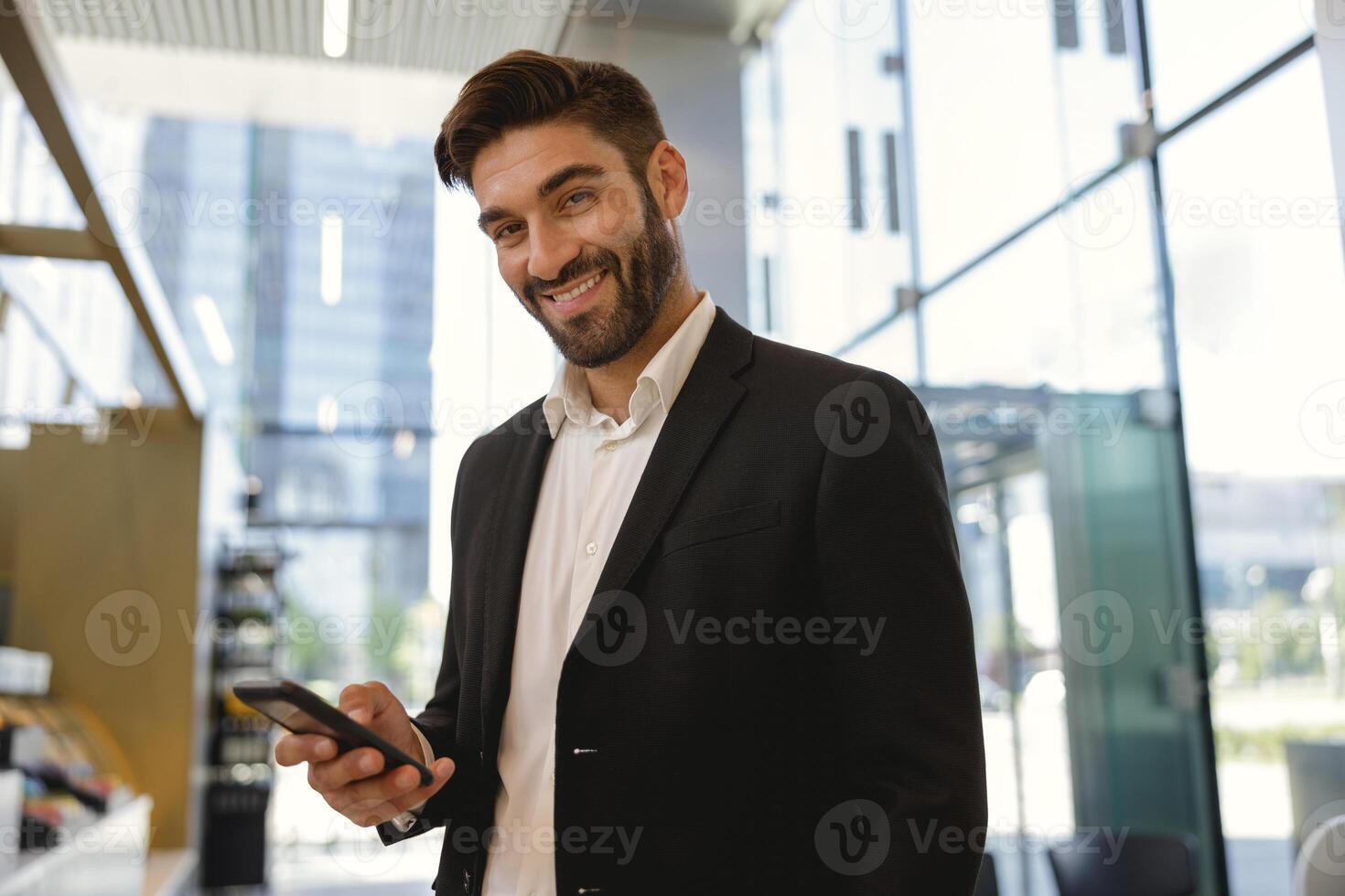 Businessman holding mobile phone and looking at camera with smile standing on office background photo