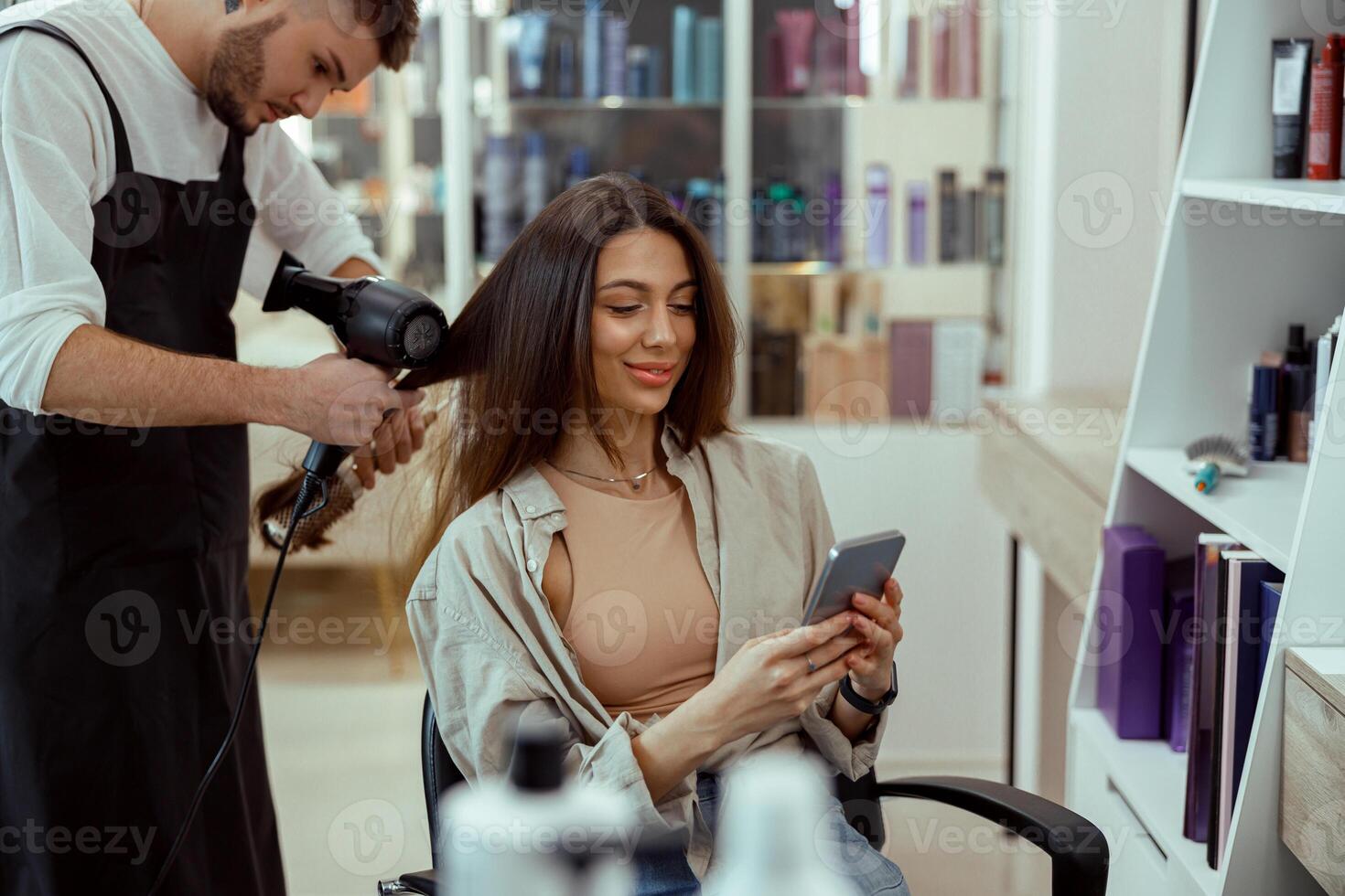 belleza salón hembra cliente utilizando su teléfono mientras estilista haciendo su pelo foto
