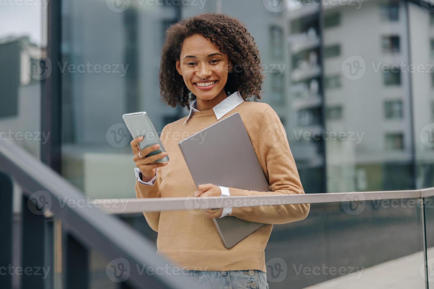 sonriente mujer gerente es utilizar teléfono mientras en pie con ordenador portátil en moderno edificio antecedentes foto