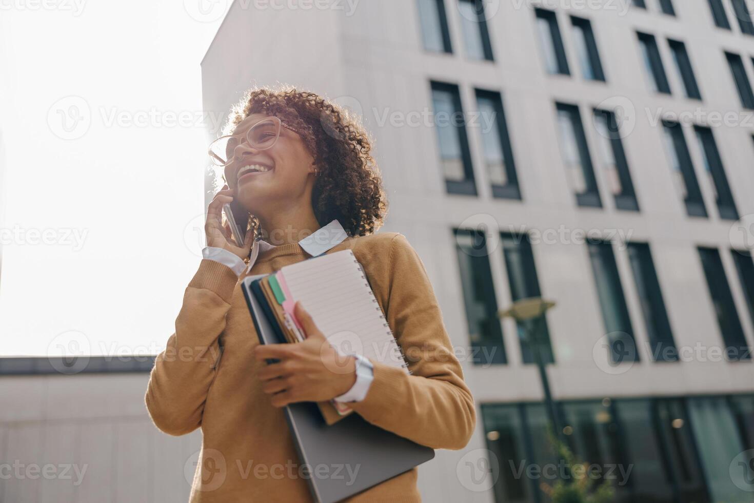 Smiling woman manager is talking phone while standing with laptop on modern building background photo