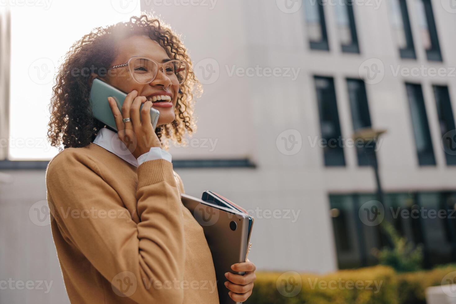 Smiling business woman is talking phone while standing with laptop on modern building background photo