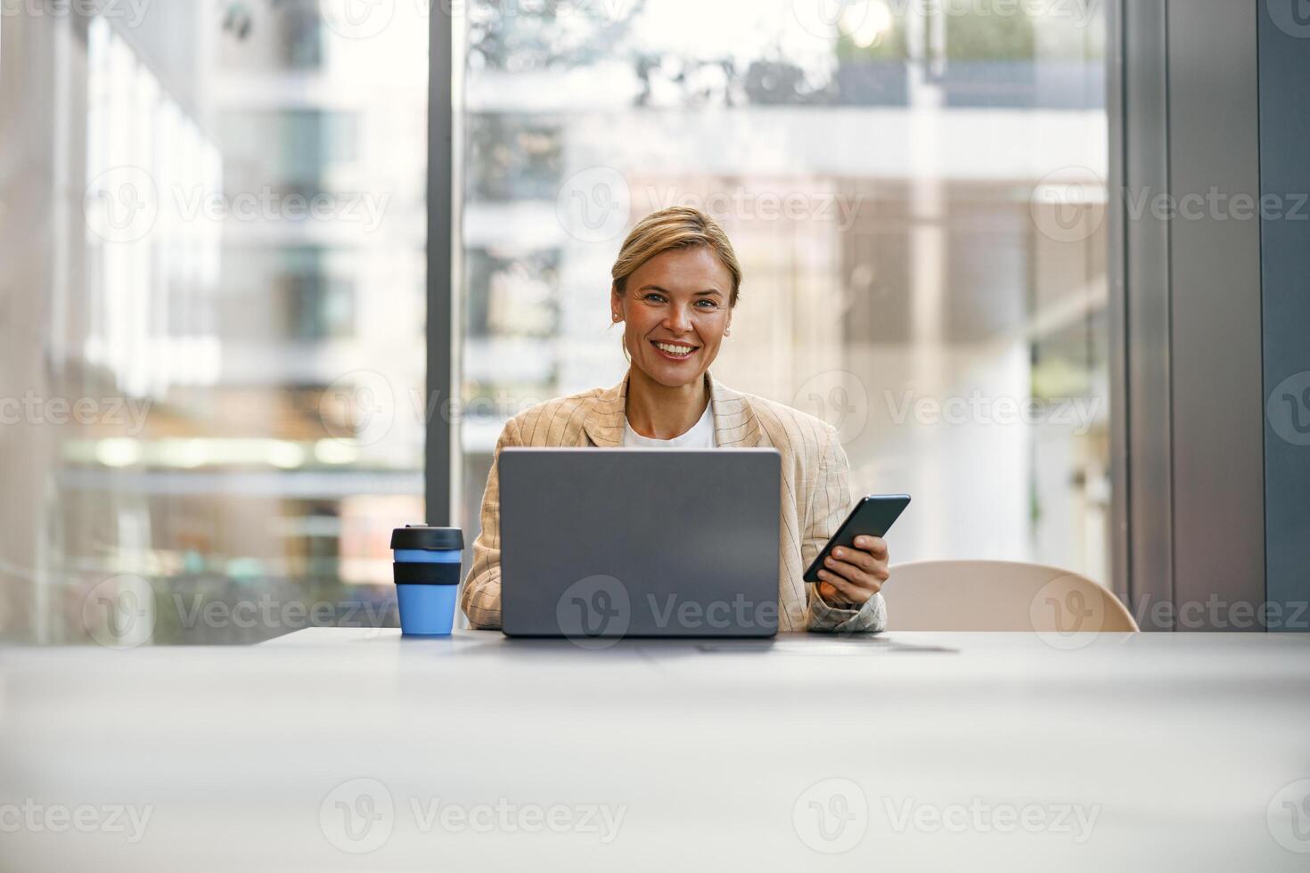alegre hembra gerente utilizar teléfono mientras sentado en moderno trabajo colaborativo y trabajando en ordenador portátil foto
