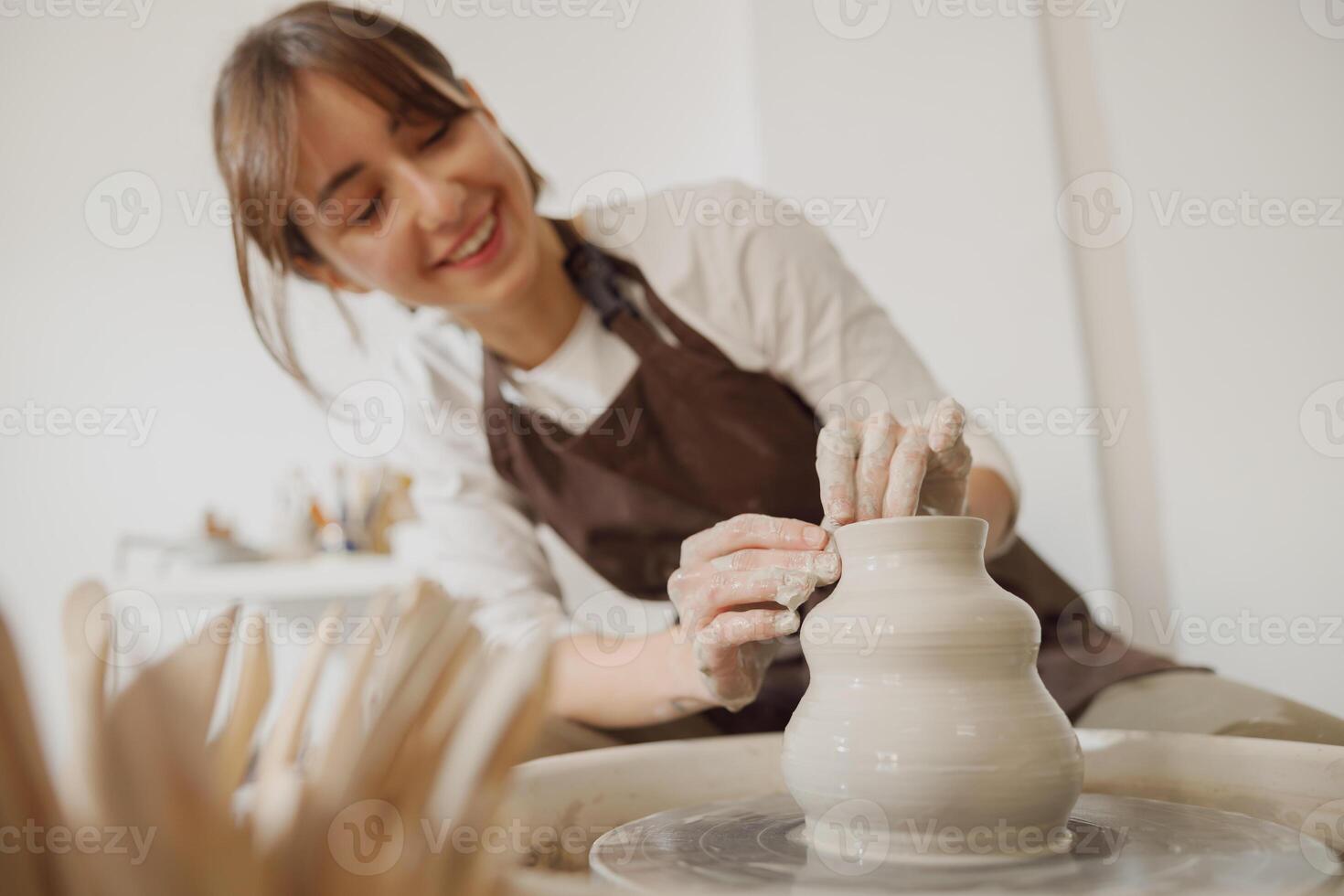 sonriente hembra artesano en delantal sentado en banco con cerámica rueda y haciendo arcilla maceta foto