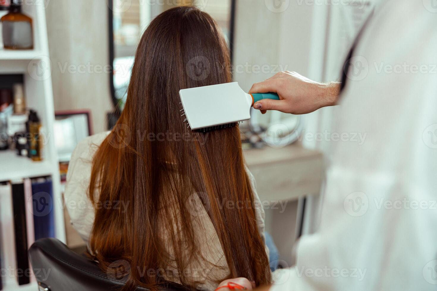 Hand of hairdresser brushing long and shiny brown hair of young woman at beauty salon photo