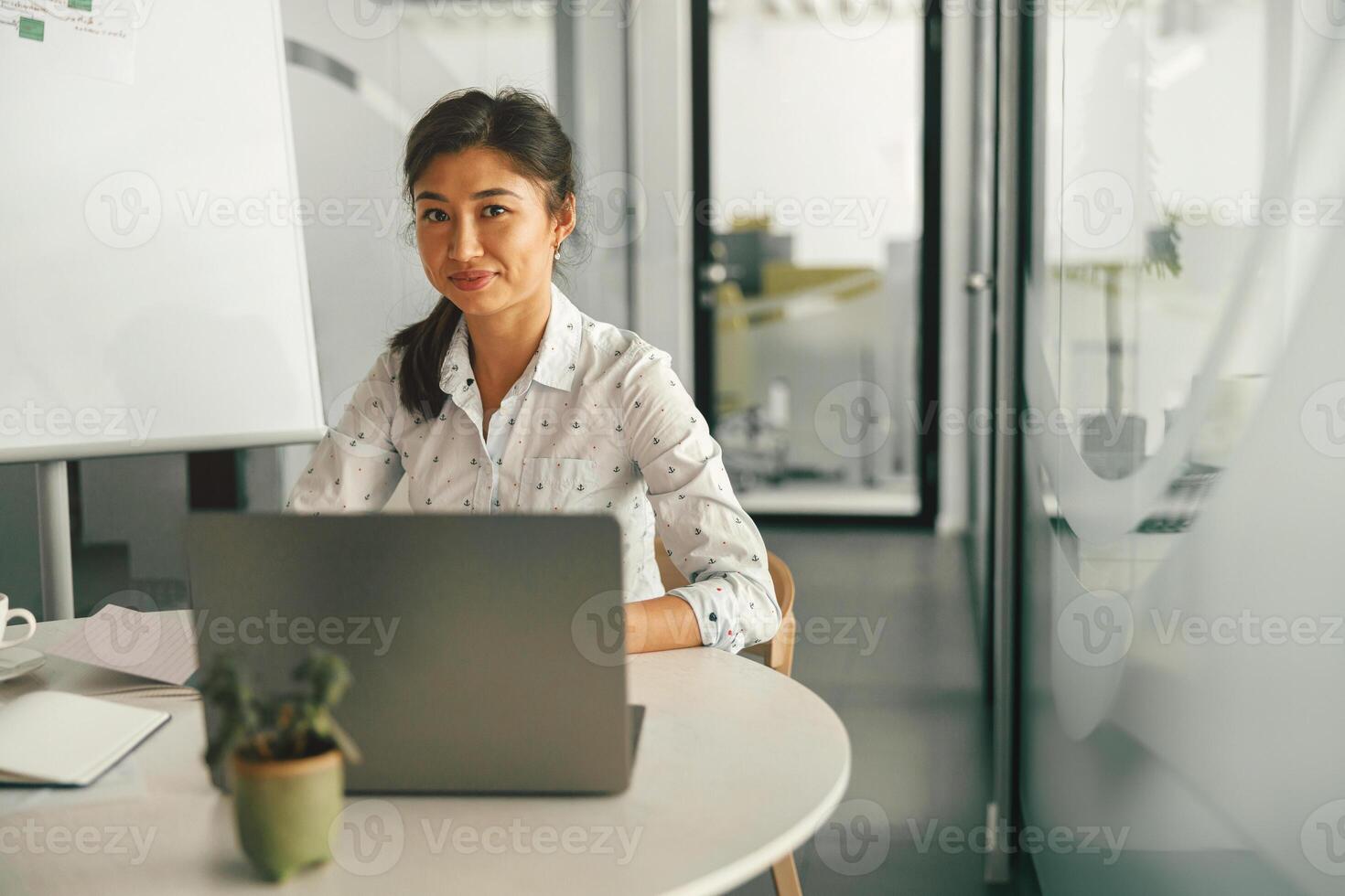 hembra gerente trabajando en ordenador portátil mientras sentado el escritorio en moderno trabajo colaborativo y mirando a cámara foto
