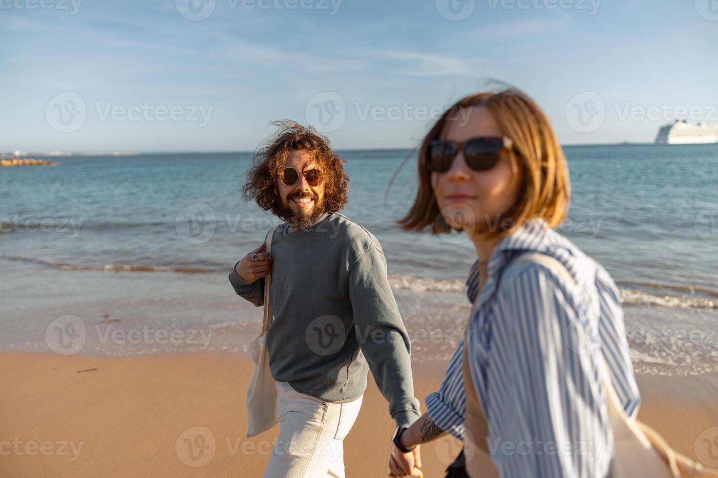 cerca arriba de romántico Pareja en amor es caminar participación manos a través de soleado playa y mirando a cámara foto