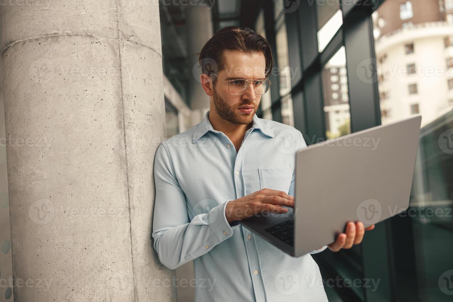 Focused male sales manager in eyeglasses working on laptop standing in office during working day photo