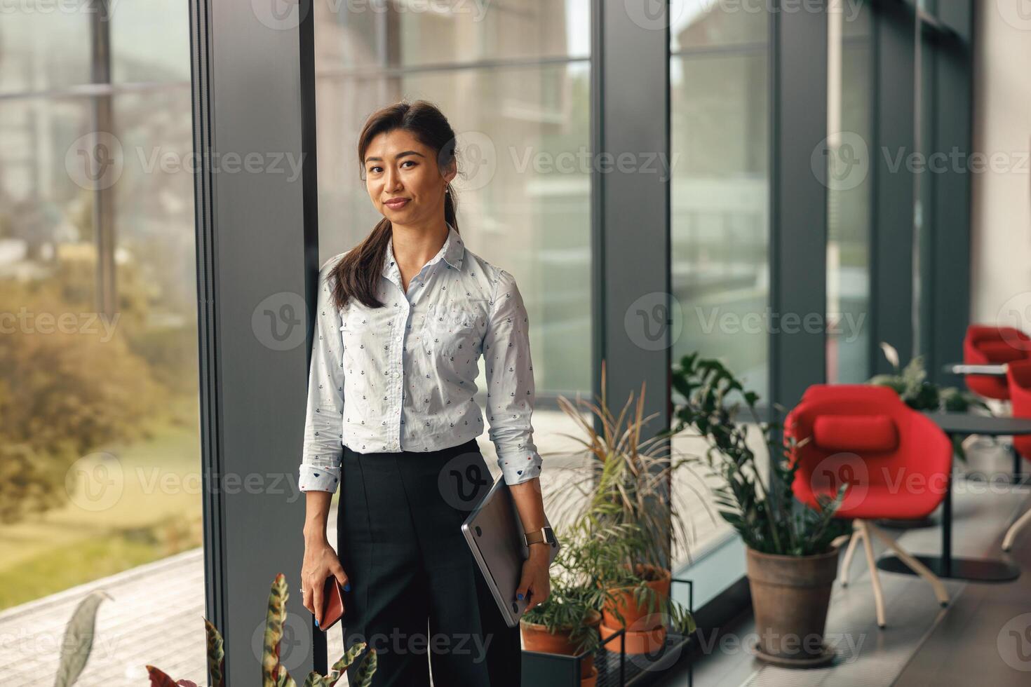 retrato de elegante negocio mujer en pie con ordenador portátil cerca oficina ventana y mirando a cámara foto