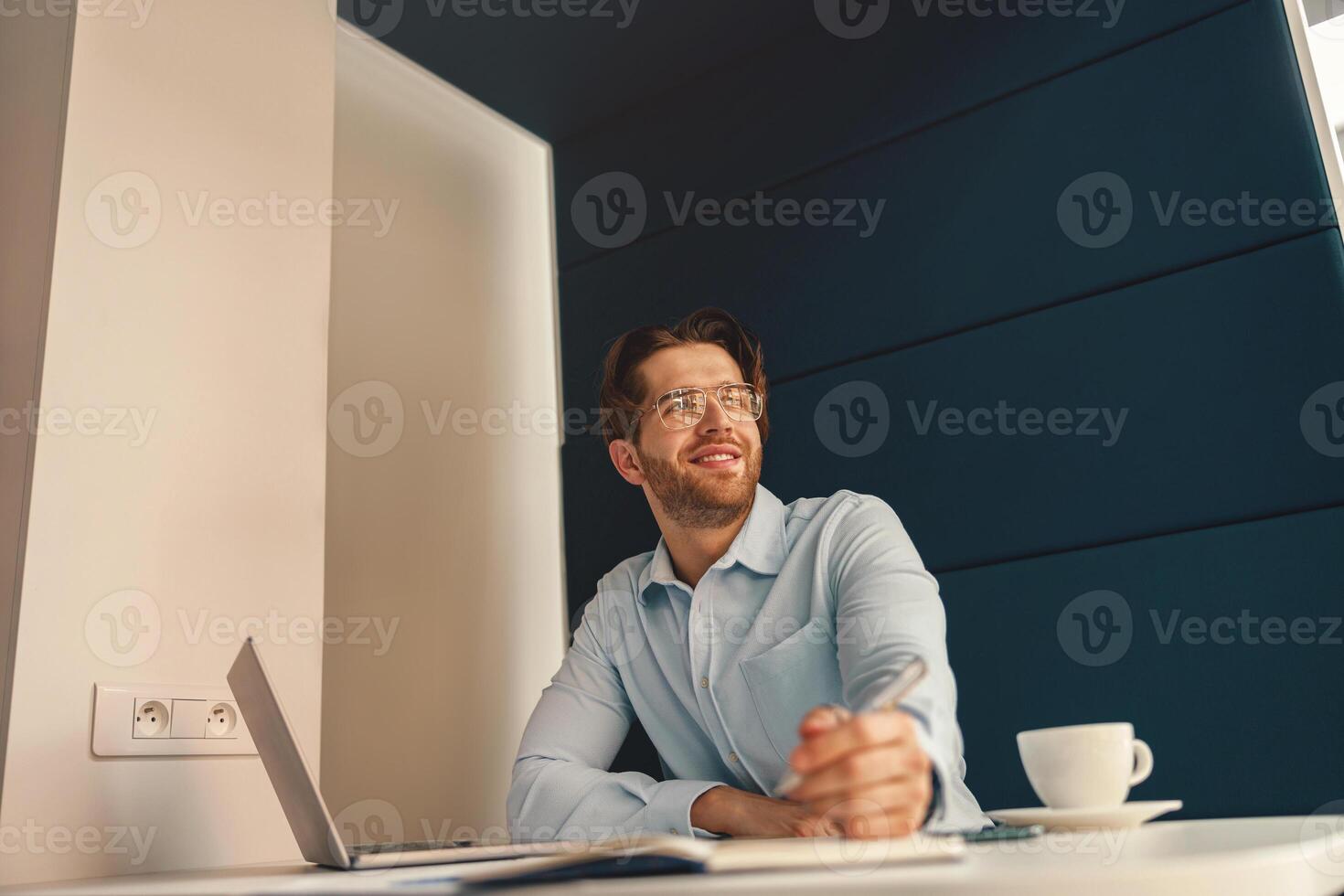 Smiling male freelancer making notes during working on laptop in coworking and looking at side photo