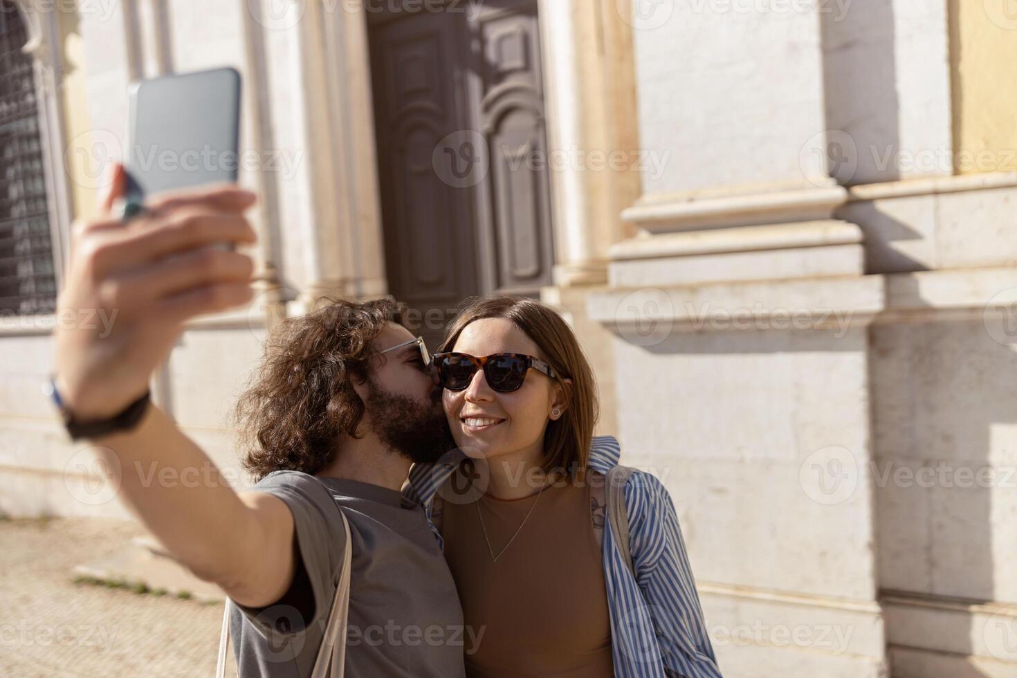 Happy tourist couple walking in city street, kissing and making selfie on phone photo