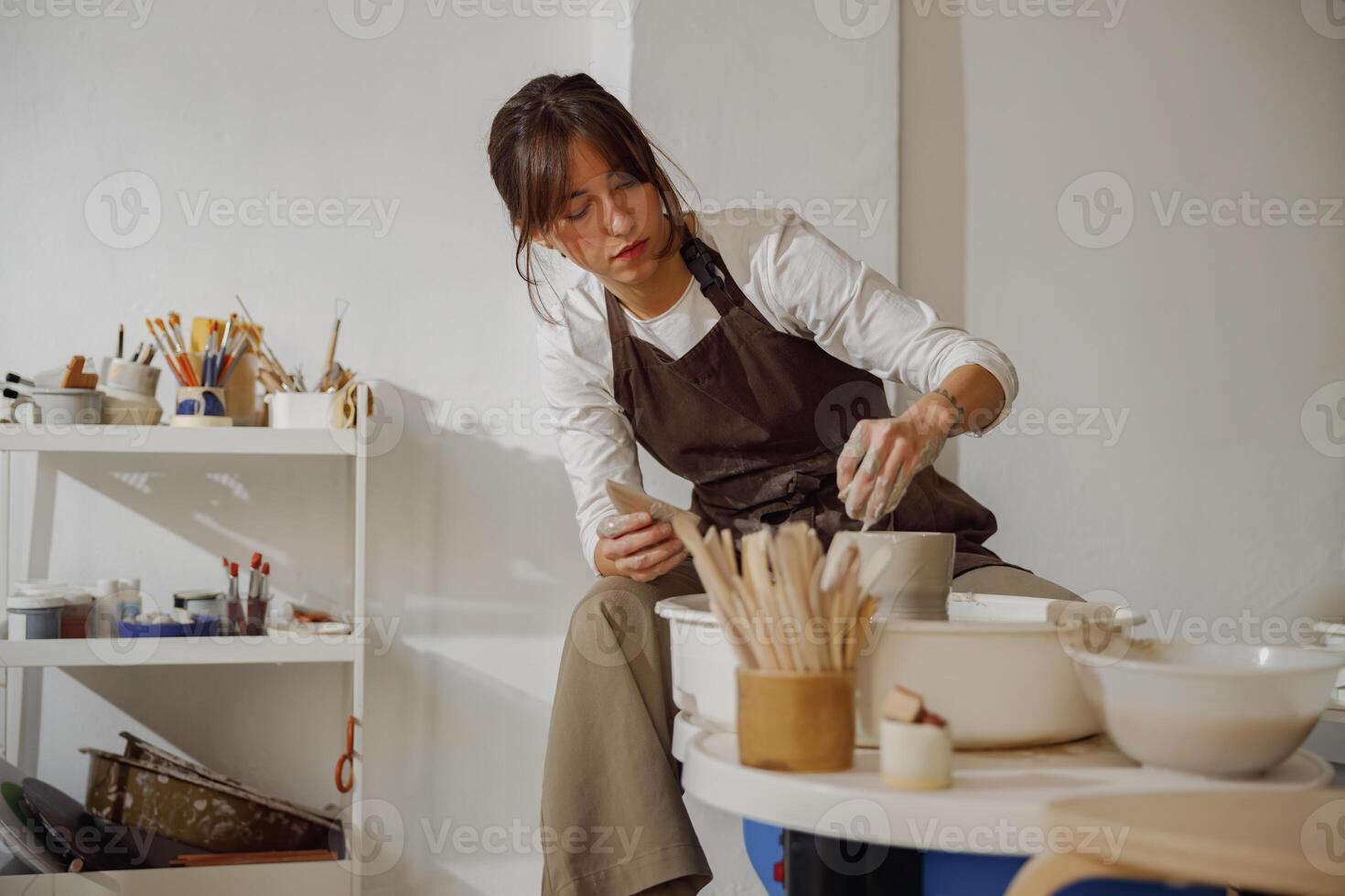 enfocado hembra artesano en delantal sentado en banco con cerámica rueda y haciendo arcilla maceta foto