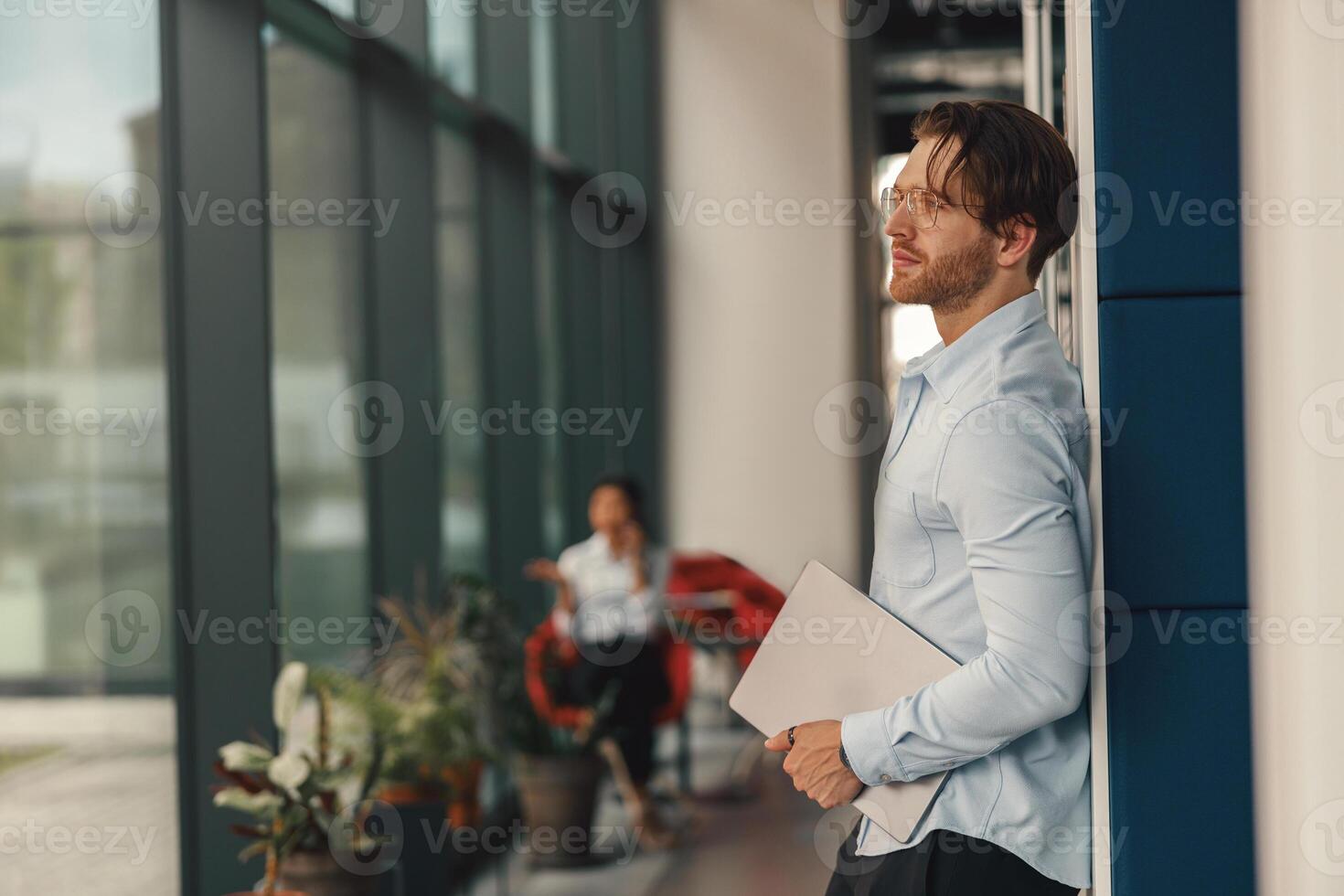 Stylish businessman with laptop standing in office hall on colleague background and looking at side photo