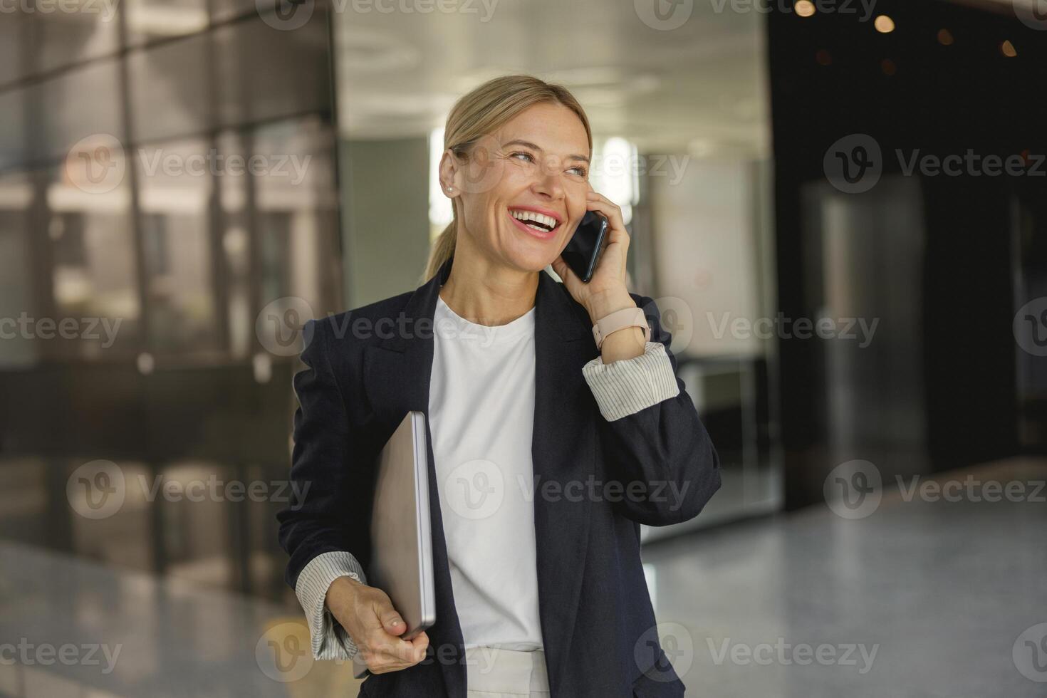 alegre mujer de negocios es hablando teléfono en pie en antecedentes de moderno oficina salón y mira lejos foto