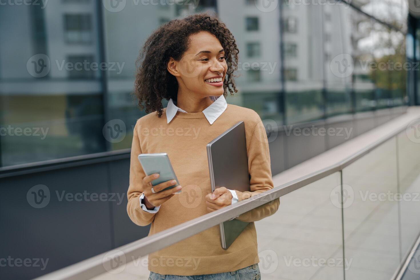 Pretty female entrepreneur is use phone while standing with laptop on modern building background photo