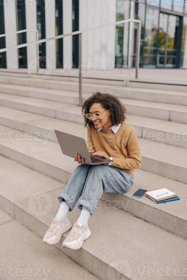 sonriente joven hembra estudiante trabajando en ordenador portátil sentado en escalera en moderno edificio antecedentes foto