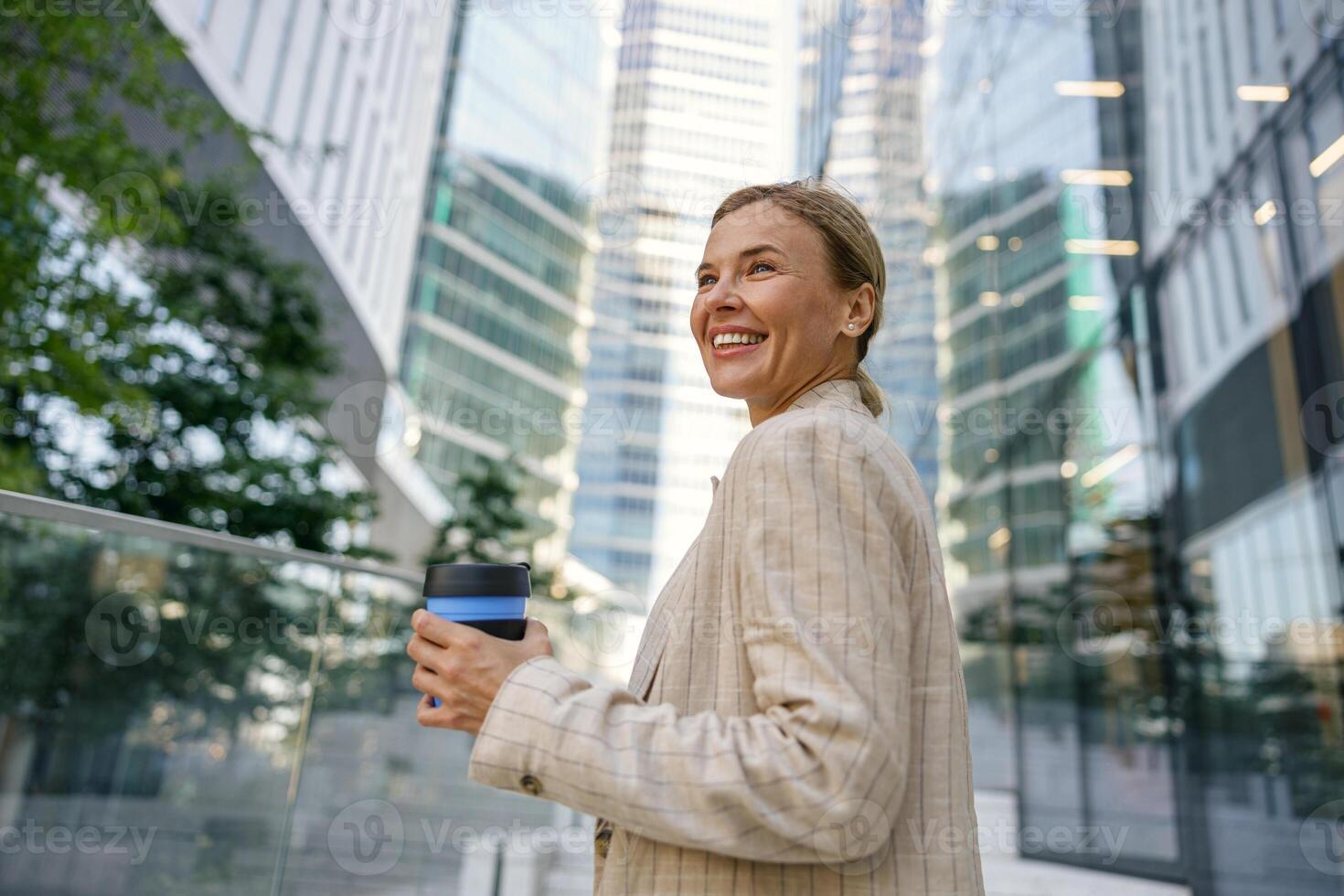 alegre negocio mujer Bebiendo café durante descanso hora cerca oficina edificio y mira lejos foto