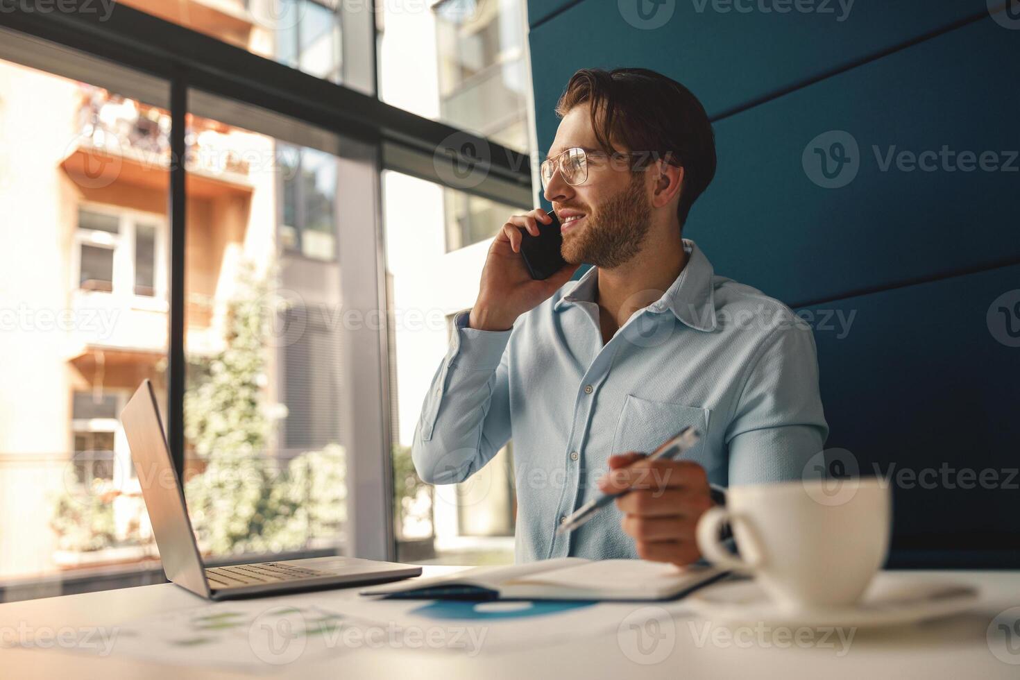 Male boss talking phone and making notes while sitting in modern office and working on laptop photo