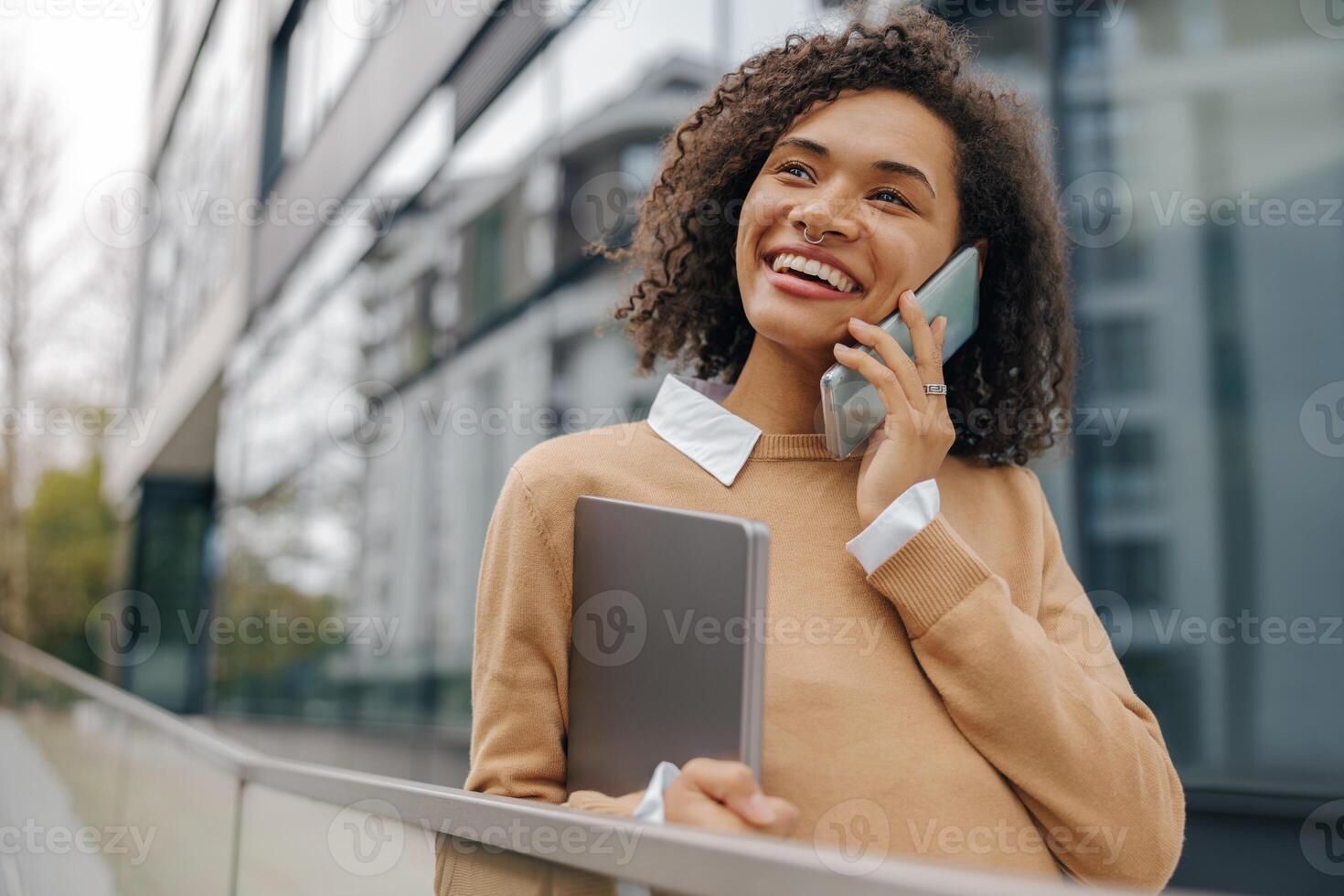 Smiling woman manager is talking phone while standing with laptop on modern building background photo