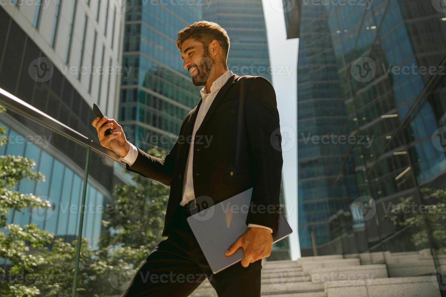 Handsome businessman using mobile phone standing with laptop on background of skyscrapers photo