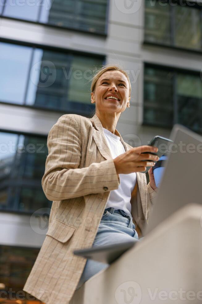 Smiling female entrepreneur use phone while working on laptop in modern office background photo