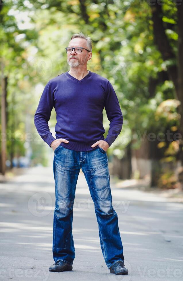 Middle age man with gray hair in park. Blue sweater. Eyeglasses. Bearded confident businessman. Blurred background. Hands in pockets. Half size photo