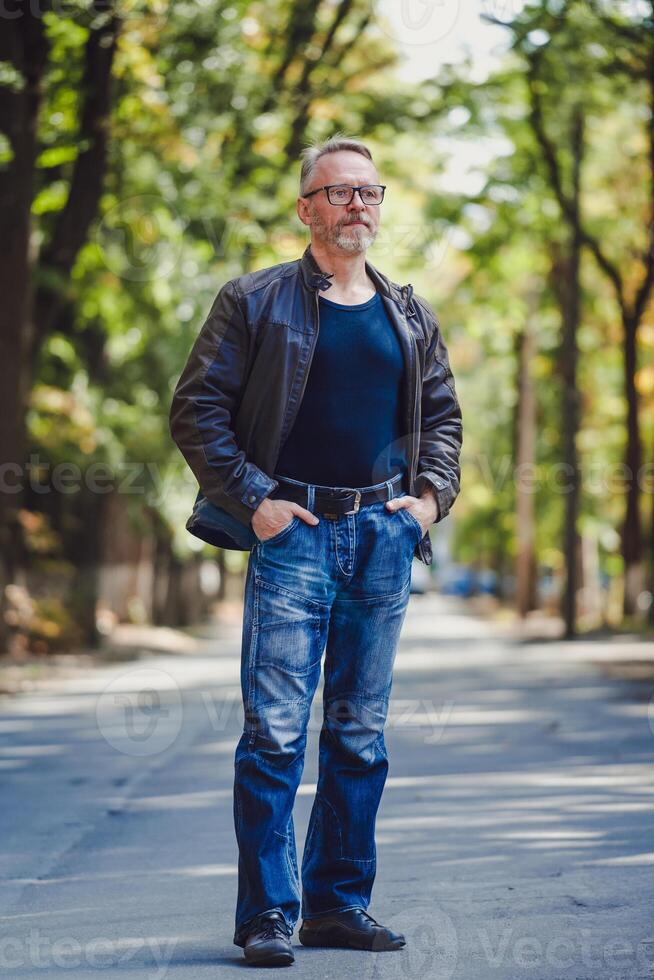 mayor hombre con gris pelo en un al aire libre ajuste. cuero chaqueta. azul suéter. los anteojos. barbado confidente empresario. borroso antecedentes. manos en bolsillos foto