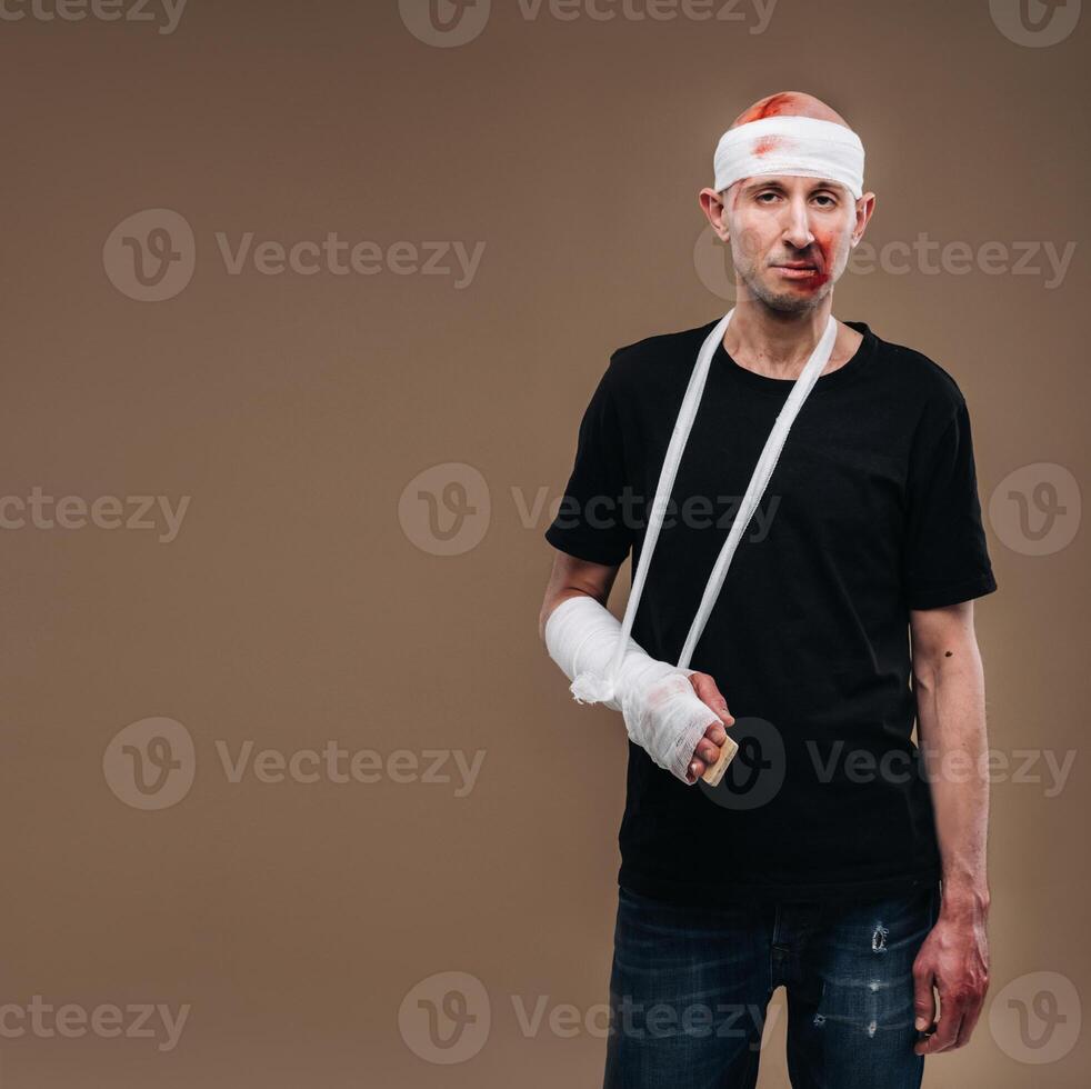 A battered man with a bandaged head and a cast on his arm stands on crutches on a gray background photo