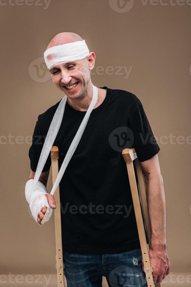 A battered man with a bandaged head and a cast on his arm stands on crutches on a gray background photo