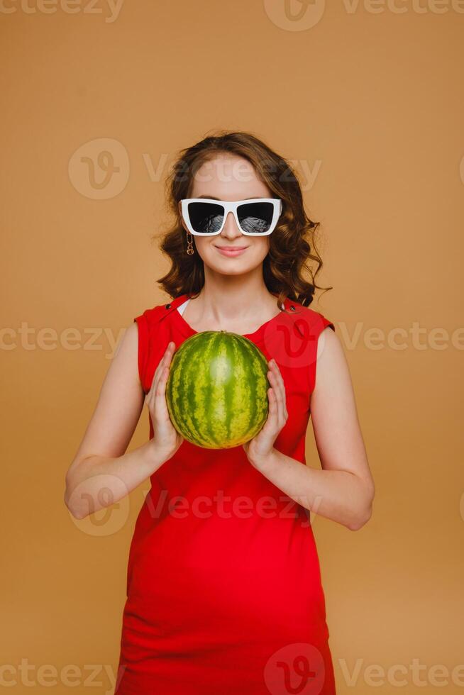 A beautiful girl in glasses and a red dress holds a watermelon in her hands photo