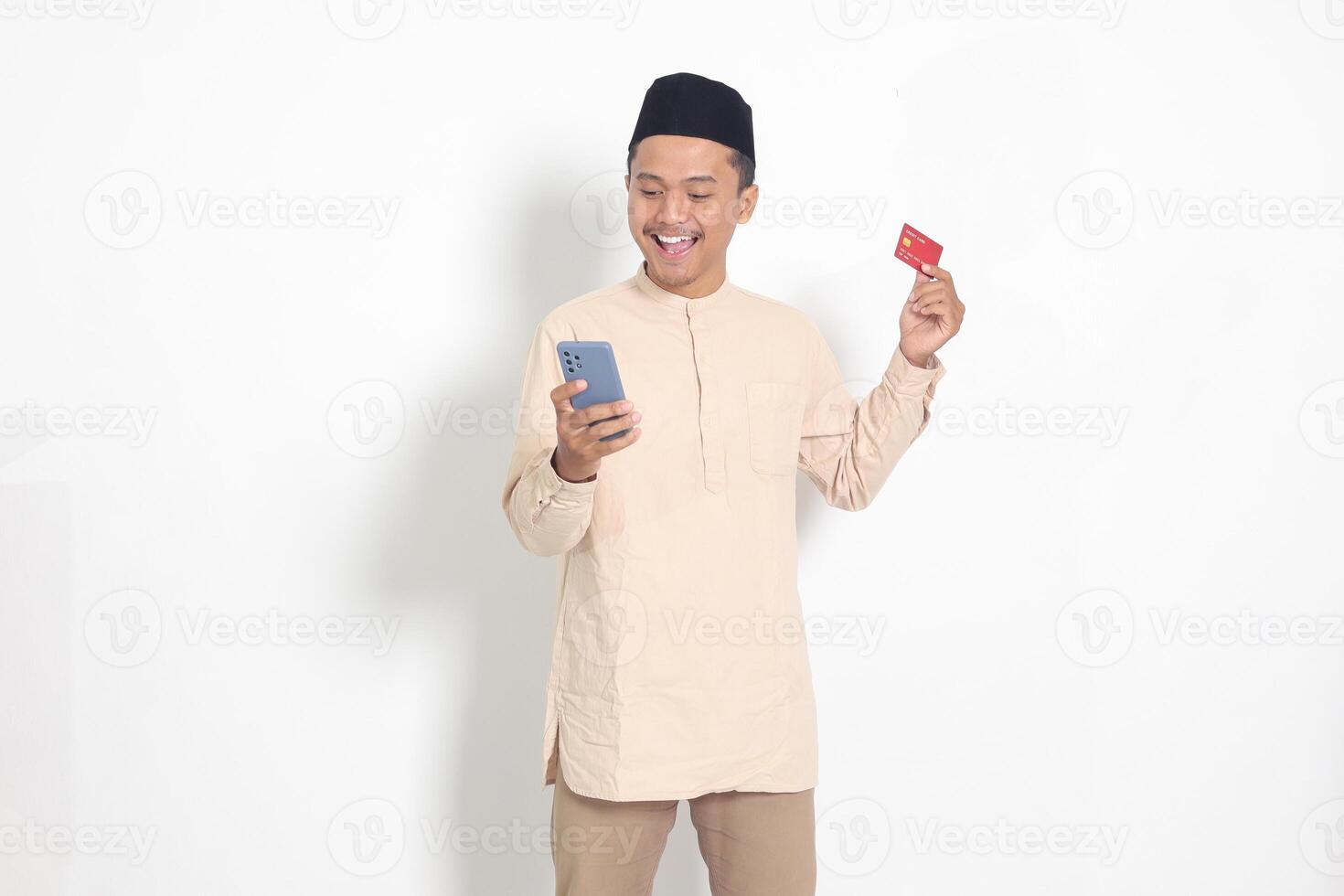 Portrait of attractive Asian muslim man in koko shirt with skullcap holding a mobile phone and presenting credit card. Isolated image on white background photo