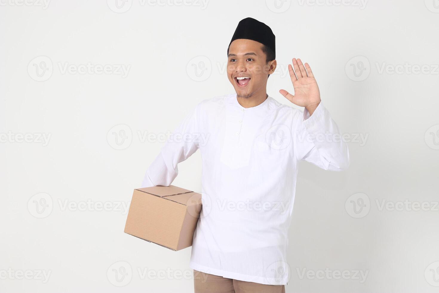 Portrait of excited Asian muslim man in koko shirt with peci carrying cardboard box while saying hi, greeting to someone. Going home for Eid Mubarak. Isolated image on white background photo