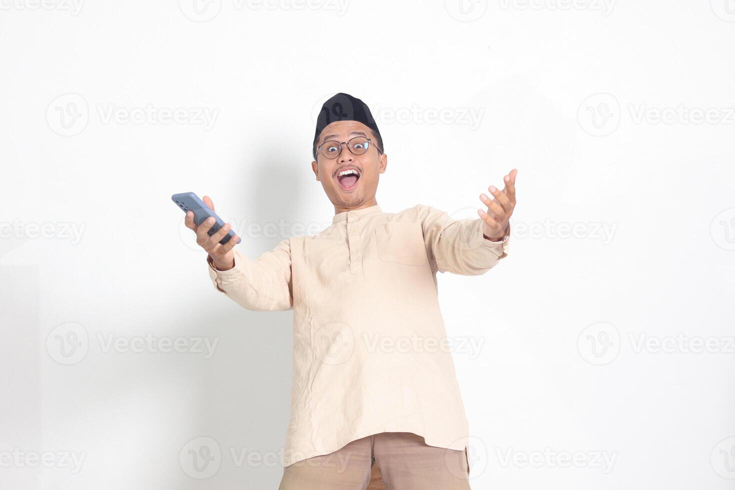 Portrait of surprised Asian muslim man in koko shirt with skullcap holding mobile phone, showing wow shocked face expression. Advertising and social media concept. Isolated image on white background photo